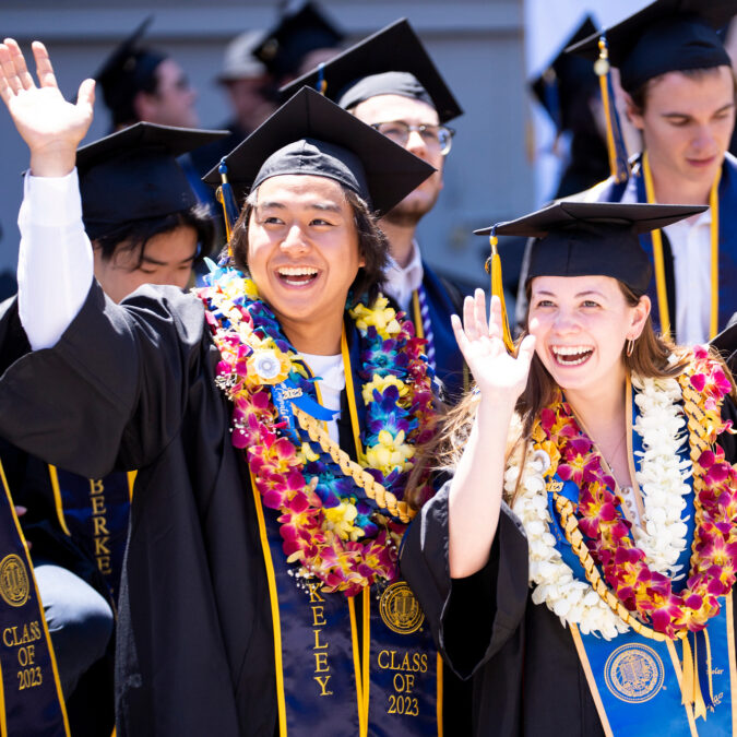 Two graduates smile and wave at commencement while wearing graduation regalia.