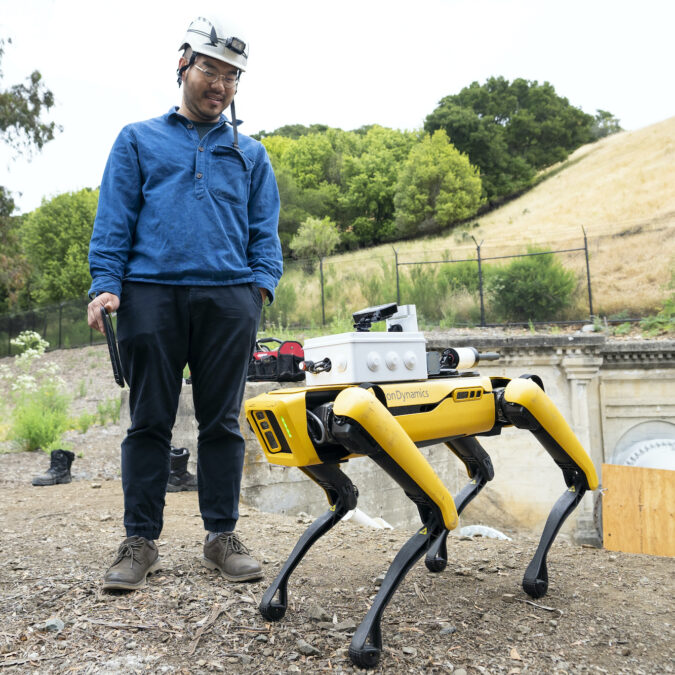 Wonjun Cha (left) in a blue shirt and white hard hat, looks down at Spot, a yellow and black quadruped robot.