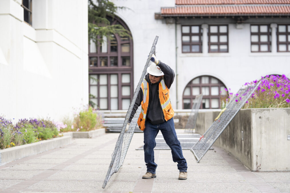 A construction worker in a hard hat and bright orange vest holds up a piece of fencing.
