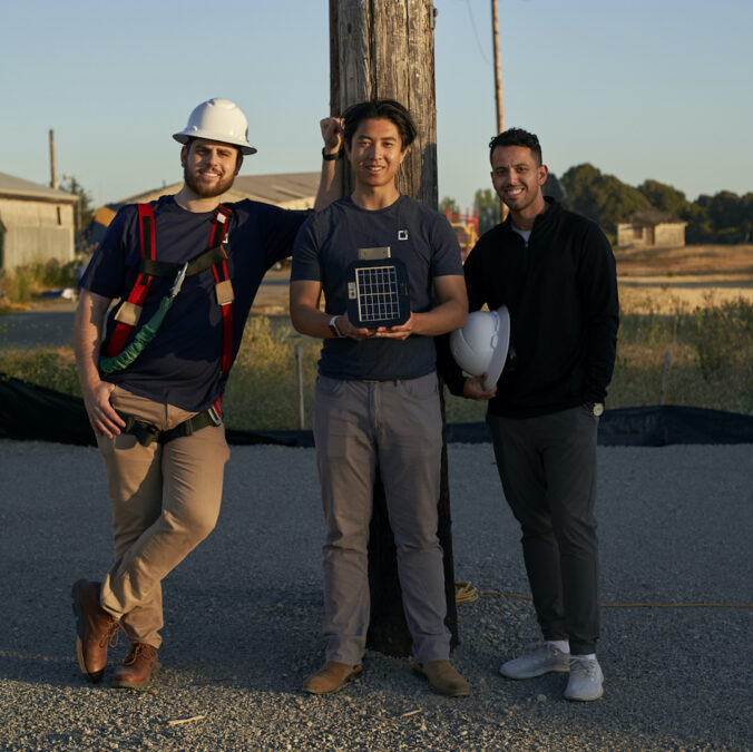 Three people pose in front of a power line pole wearing hardhats and holding a rectangular device.