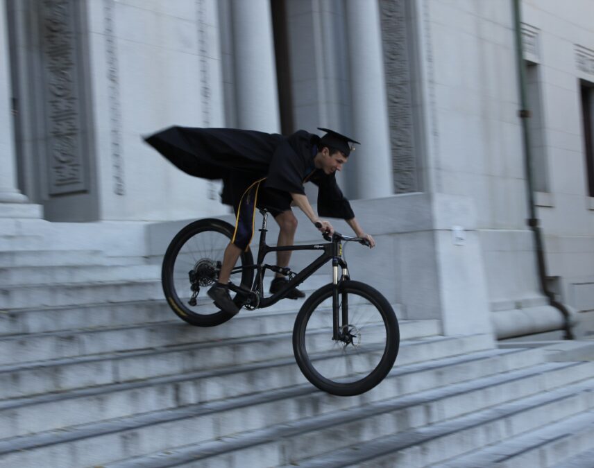 Ziven Posner rides a bicycle down the front steps of the Hearst Memorial Mining Building while wearing graduation regalia that billows from behind.