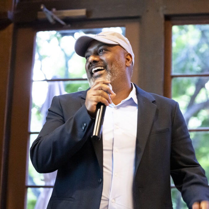 A man in a baseball cap and blazer smiles while holding a microphone.