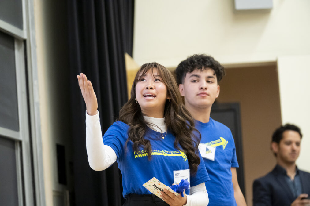 A student in a blue "Pre-Engineering Program" shirt gestures to the top-left corner while giving a presentation.