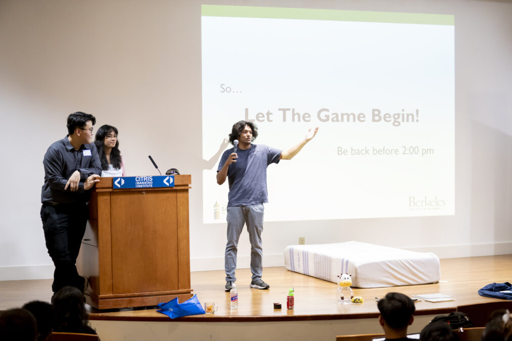 Three students give a presentation in front of a projector slide that reads, "Let The Game Begin!"