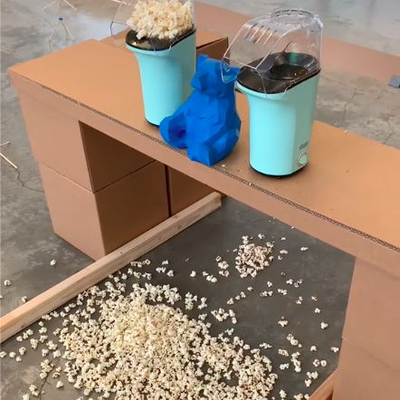 A blue bear figurine sits on a desk flanked by two popcorn makers, with popcorn strewn on the floor below.