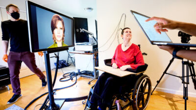Research participant, sitting in a wheelchair, is connected by wires to a computer and monitor that displays an avatar. A research coordinator can be seen walking behind the monitor, and in the foreground, someone is holding a laptop computer.