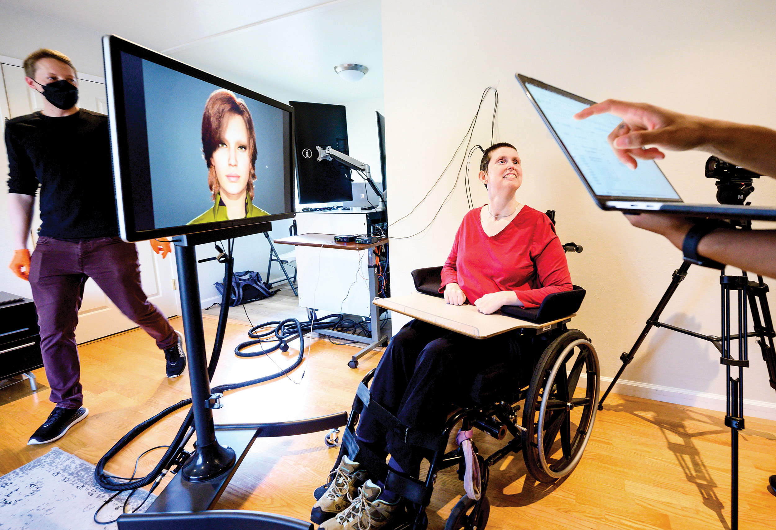 Ann, in a wheelchair, is connected to computers that translate her brain signals into the speech and facial movements of an avatar. At left is UCSF clinical research coordinator Max Dougherty.