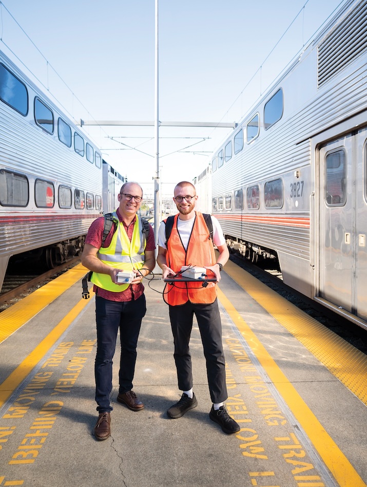 Civil and environmental engineering professor Joshua Apte and postdoc Sam Cliff smiling at the camera, standing on a Caltrains platform between two trains (Photo by Adam Lau/Berkeley Engineering)