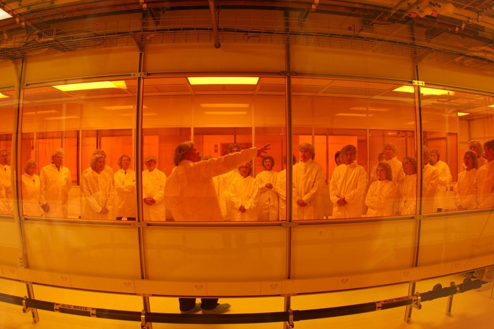 A group of people in lab coats and hairnets listening to a man in lab safety gear pointing to his right