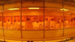Shot of more than 20 people in a room wearing clean suits. The people are listening to a person talking and pointing to the right.