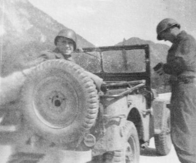 Esta foto da Segunda Guerra Mundial mostra George Leitmann sentado em um jipe, vestindo um capacete, olhando para a câmera enquanto o motorista está de pé para o lado.