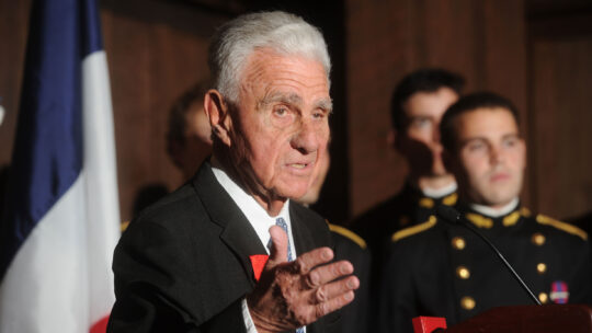 George Leitmann, wearing a suit and tie, gestures at the lectern. In the background are French cadets in uniform.