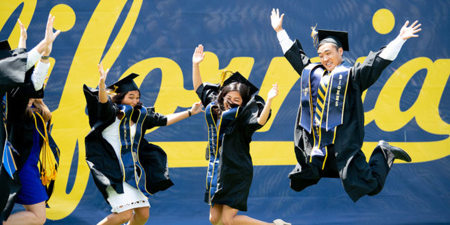 Graduates jumping in caps and gowns in front of a large script California banner